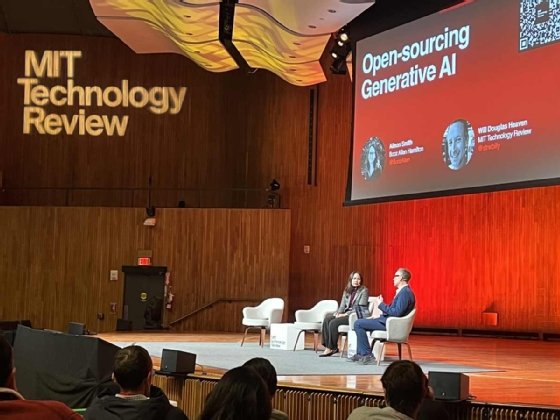 Two people sit on an auditorium stage with a slide titled 'Open Sourcing Generative AI' and 'MIT Technology Review' projected on the wall behind them.