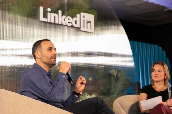 A man is speaking into a microphone on a LinkedIn stage while a woman with a microphone and notes is listening.